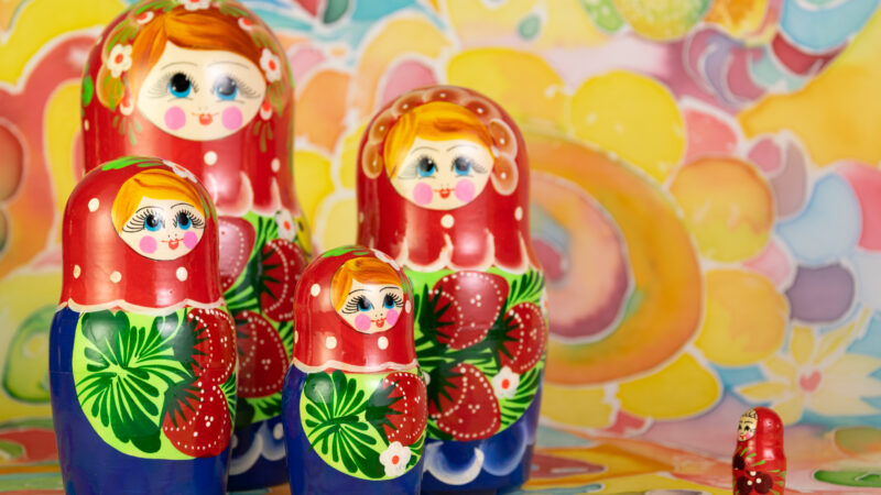 Eine russische Matroschka aus 5 Puppen vor einem Seidentuch mit vielfarbigen Mustern, Figuren und einer Spirale im Zentrum. 4 Puppen stehen links, die kleinste steht rechts - ihr Gesicht ist den anderen zugewandt.