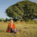 Doktor Christine Wallner sitzt vor einem großen Baum, dem sogenannten “Africa Amini Alama-Baum” im Zentrum von Momella, Tansania