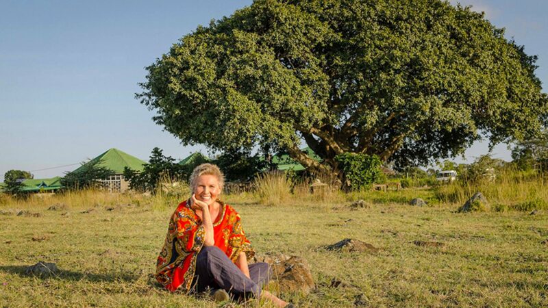 Doktor Christine Wallner sitzt vor einem großen Baum, dem sogenannten “Africa Amini Alama-Baum” im Zentrum von Momella, Tansania