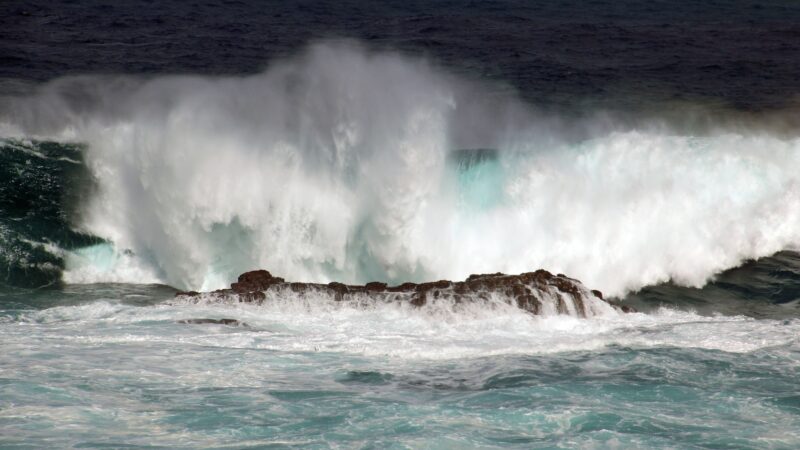 Man blickt von einer Küste hinaus auf das stürmisches Meer. Im Zentrum des Bildes liegt ein breiter, eher flacher Felsen, an welchem sich die Wellen brechen.