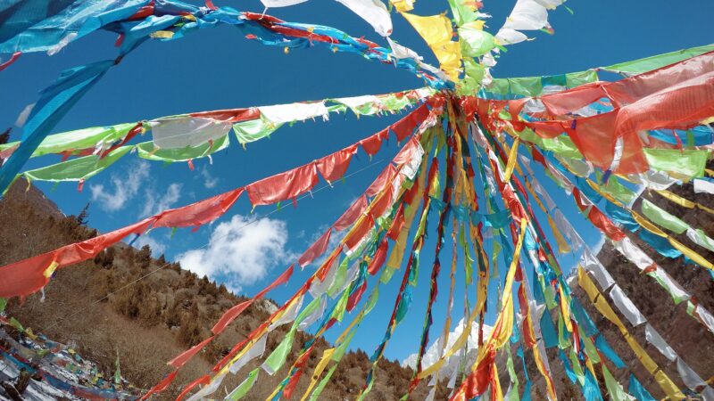 Von unten mit Blick auf den Himmel sieht man viele bunte Gebetsfahnen, die sternförmig zusammengebunden sind.