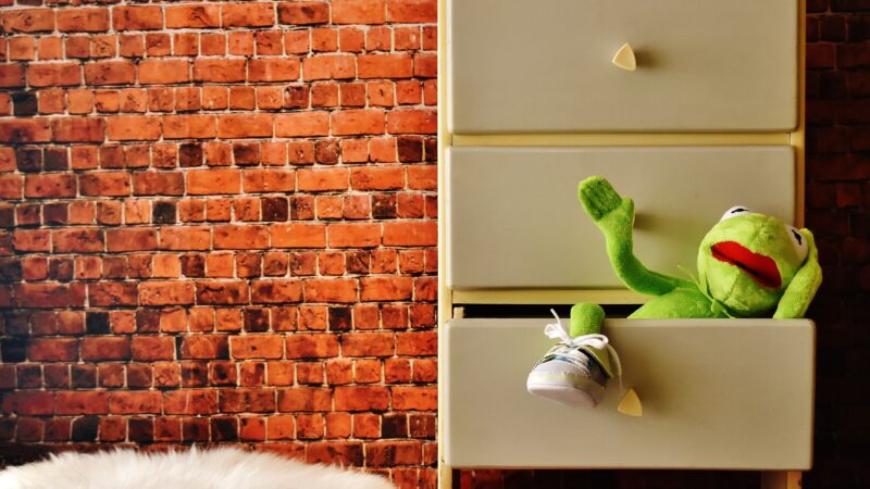 Vor einer Ziegelwand steht ein Kästchen mit drei Schubladen. Aus der untersten heraus winkt uns die Puppe "Kermit der Frosch" zu.