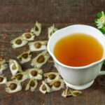 Links neben einer Tasse mit Tee liegen die Kapselfrüchte der Moringa-Pflanze, rechts davon ihre Blätter und Blüten.