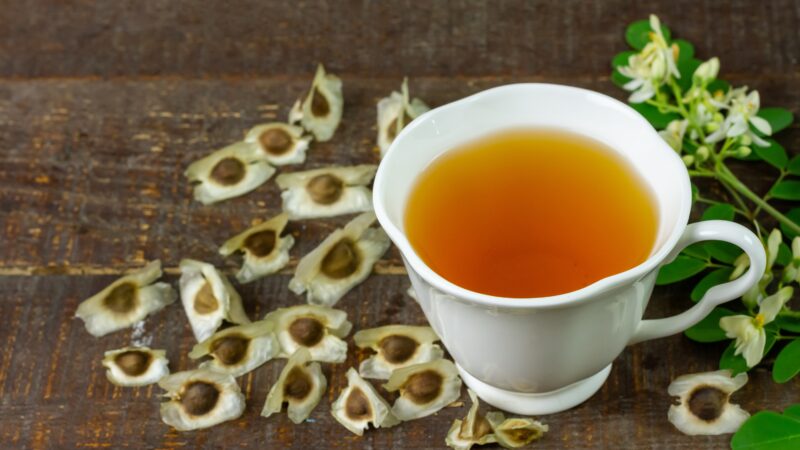 Links neben einer Tasse mit Tee liegen die Kapselfrüchte der Moringa-Pflanze, rechts davon ihre Blätter und Blüten.