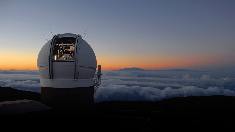 Kuppel des PAN-STARRS Teleskops des Haleakala Observatoriums auf Hawaii. Im Hintergrund ein Nebelmeer und der freie Himmel bei Dämmerung.