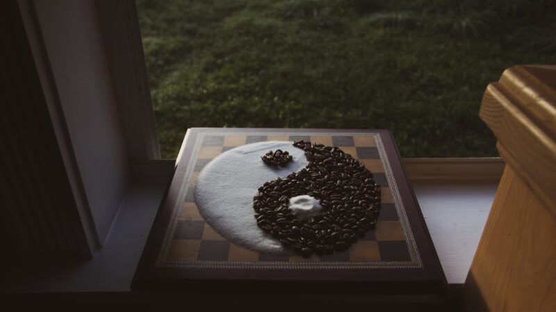 Auf einem Fensterbrett liegt ein Schachbrett. Darauf ist aus Zucker und Kaffeebohnen ein Yin-und-Yang-Symbol aufgestreut. Jenseits des Fensters sieht man ins Grüne.