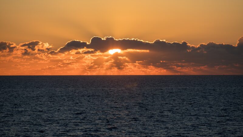 Sonnenaufgang über dem Ozean. Knapp über dem Horizont befindet sich eine Wolkenbank. Die Sonne scheint durch einen Spalt hindurch.