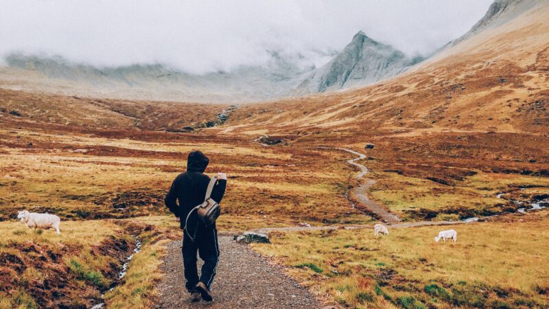 Ein Wanderer auf einem Pfad, der durch eine Hochebene führt. Im Hintergrund hohe Berge.
