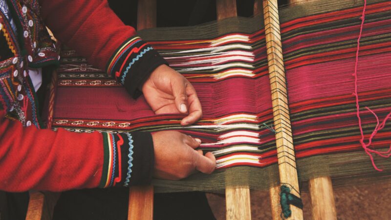 Ein Gewebe - möglicherweise ein Mantel oder Poncho - in der Entstehung. Das Gewebe ist bunt, vorallem mit viel rot, aber auch weiß, grün, orange und schwarz sind vertreten. Von links kommend sieht man die Hände der Person, welche das Gewebe herstellt.