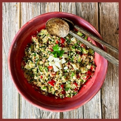 In einer runden roten Schüssel ist ein Salat von Couscous, Tomate und Gurke angerichtet. Darüber wurden Petersilie und Schafkäse gestreut. 