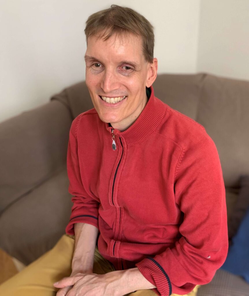 Energetiker Clemens Pistauer sitzt auf einem Sofa in seiner Praxis und lächelt in die Kamera. Er hat dunkelblonde Haare und trägt eine rote Weste.