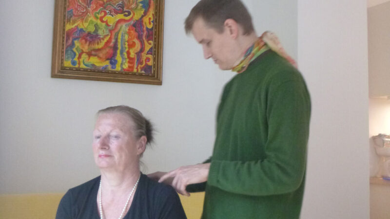 Clemens Pistauer behandelt eine Klientin energetisch. Er steht hinter ihr und berührt sie mit zwei Fingern leicht an den Schultern.