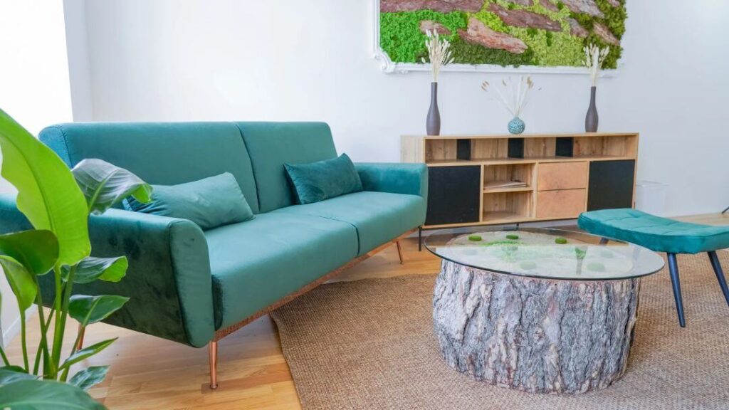 Heller Praxisraum mit Parkettboden. Links steht ein türkisfarbenes Sofa, daneben befindet sich ein abgeschnittener Baumstamm als Beistelltisch. An der Wand hängt ein Bild aus Moos.